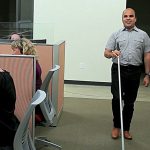a man is walking with a white cane between cubicles of people working