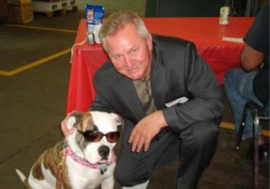 Jim Kerlin poses next to a brown and white dog wearing sunglasses.