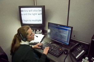 A woman is sitting at a computer and wearing a phone headset. To her left is a screen magnifier.
