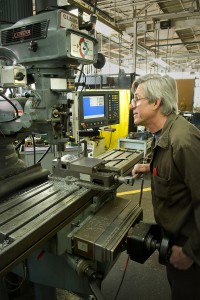 A man with heavy glasses is looking at a drill press.