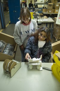 A woman sits at a table putting some parts together. Another woman watches.