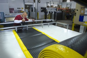 A man is at the far end of a work table with a roll of floor mat material.