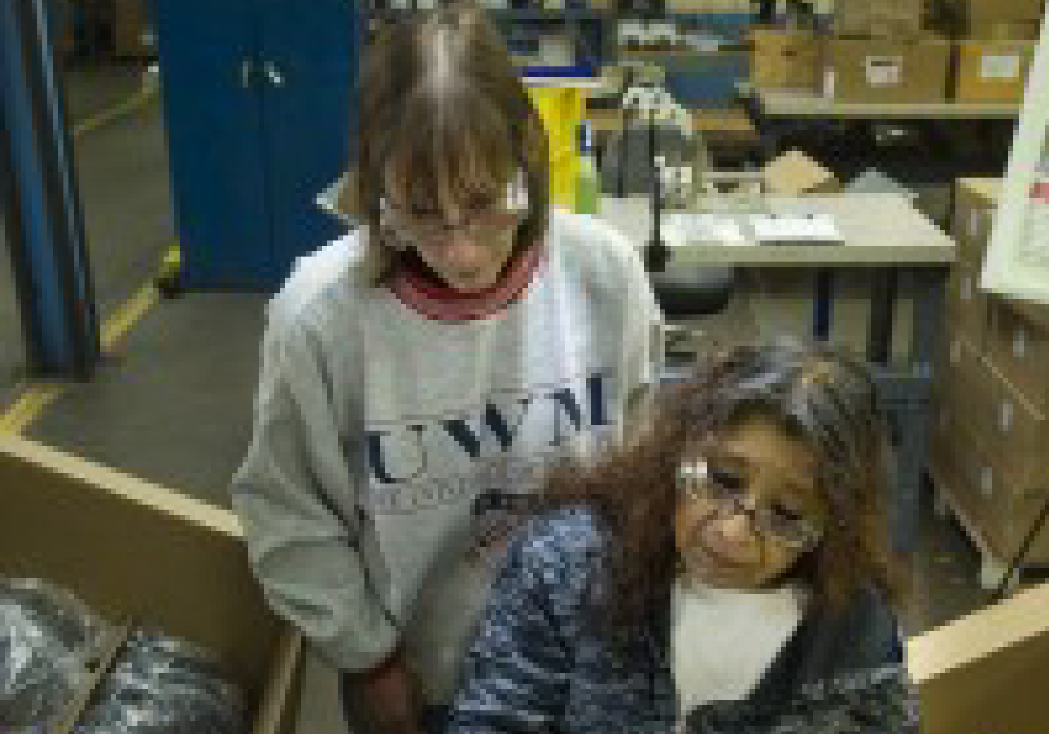 A woman sits at a table putting some parts together. Another woman watches.