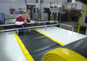 A man is at the far end of a work table with a roll of floor mat material.