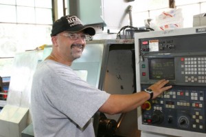 A color photograph of a man with safety glasses on working with a CNC machine.