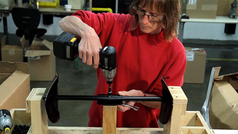 A woman is holding a drill on a piece of metal that is mounted on a workbench. She is wearing glasses and has on a red sweatshirt.