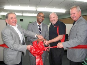 Four men are smiling holding a large red ribbon and a pair of scissors.