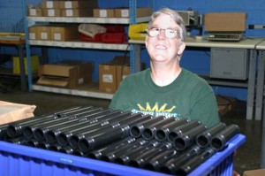 A woman with strong prescription glasses is smiling. She is in a blue room that has work benches. In front of her is a large blue, plastic bin full of parts. They are black cyclinders about 1" wide and 4" long.