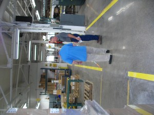 The background is a large manufacturing space. A man in a bright blue polo has his back to the camera. Another man is standing off to his right, in front of him and they are talking. The man with his back to the camera is using a cane. The yellow line on the floor is actually a raised piece of metal.