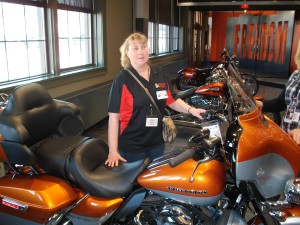 A with a Beyond Vision shirt is standing next to a Harley Davidson motorcycle. She is smilng and has a ponytail in her blond hair.