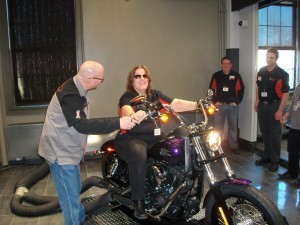 A woman wearing black jeans and a Beyond Vision shirt is sitting on a sparkly, purple Harley Davidson motorcycle. She has shoulder length brown hair and is wearing sunglasses. A man is standing next to the motorcycle showing her the levers.