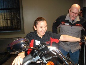 A man with a Harley Davidson shirt is standing next to a stationary Harley Davidson motorcycle. A cute young woman is smiling excitedly as she sits on the motorcycle and is reaching for the handlebars.