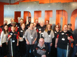 A room of people stand in two rows. Some are wearing regular clothes. Others are wearing Beyond Vision logo shirts. Several people are holding white canes. Everyone is smiling brightly.
