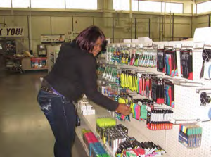 A young, african american woman is wearing jeans and a hoodie. She is in a store with office supplies. She is restocking a row of markers.