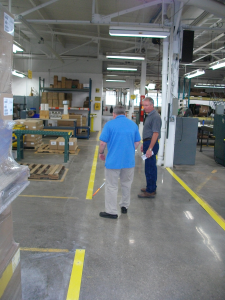 The background is a large manufacturing space. A man in a bright blue polo has his back to the camera. Another man is standing off to his right, in front of him and they are talking. The man with his back to the camera is using a cane. The yellow line on the floor is actually a raised piece of metal.