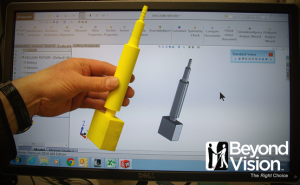 A man's hand is holding a piece of yellow plastic that is similar to a marker with a block at the end of it. In the background is a computer screen with the CAD design drawing of the item.