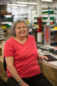 A woman who is blind smiles jokingly at the camera. She has shoulder length hair that is wavy and is a mix of grey and white. She is wearing a pink blouse and she has on a silver bracelet with a piece of turquoise on it. She is at a workstation and there is a sign that says SKILCRAFT behind her.
