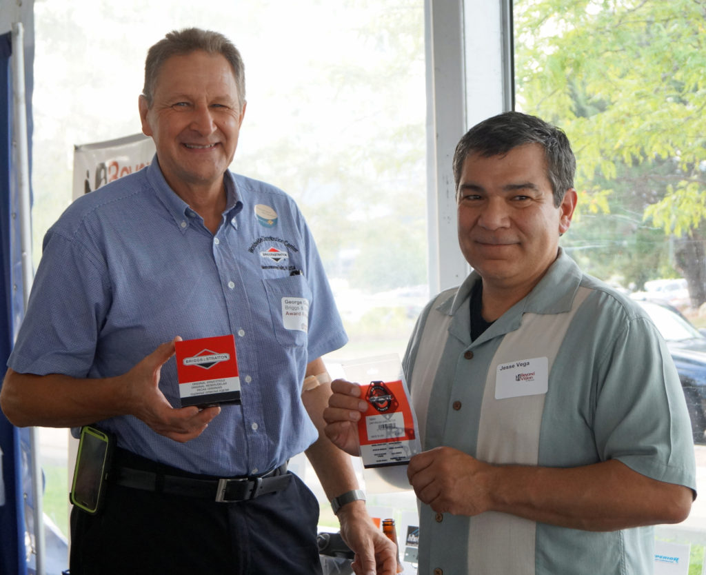 Two men standing by a table are holding Briggs & Stratton parts and smiling.