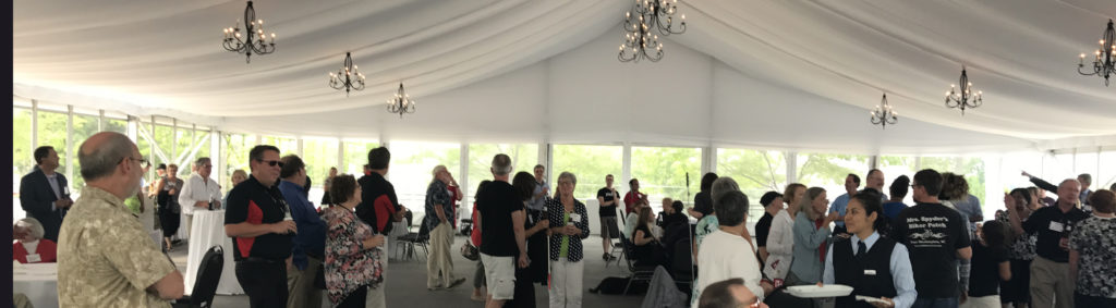 Under a tall white tent with chandeliers from the ceiling, a large crowd of people are talking, eating and drinking.