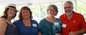 Four people stand in a row smiling at the camera. The lady on the left is wearing a straw hat with a black tank top that has white dots. She has shoulder length red hair. The next lady is wearing a blue top and also has shoulder length red hair. The couple to the right are older and both have grey and white hair and are wearing glasses. She has a blue top and he is wearing a red polo.