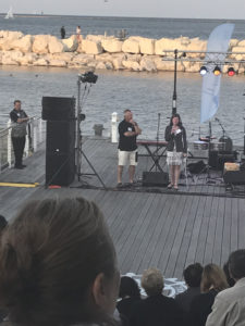 A stage at the edge of Lake Michigan. Sarah Zellmer stands at the microphone talking and JIm Kerlin stands listening as she speaks to the audience.