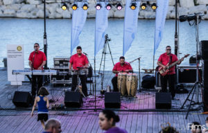 Los Ciegos del Barrio playing at LIVE at the lakefront. The stage is a pier and the water is behind them. They are all wearing red shirts and black pants. There is a man on a keyboard, a man with snare drums, a man on bongos and a man playing a guitar.