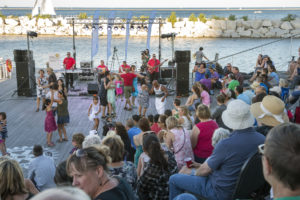A crowd of people sit facing a lower stage. Los Ciegos del Barrio are playing music and people are dancing in pairs.