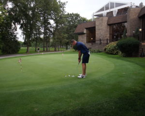 A putting green in front of a clubhouse. A man in shorts and a polo is lining up a shot.