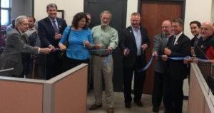 A row of ten people are holding a blue ribbon and Jim Kerlin is holding some huge scissors to cut it!