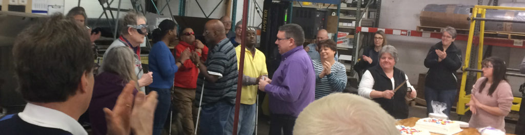 An area of a warehouse is crowded with many people around a worktable. They are clapping and smiling as a tall man stands in the center of the crowd.