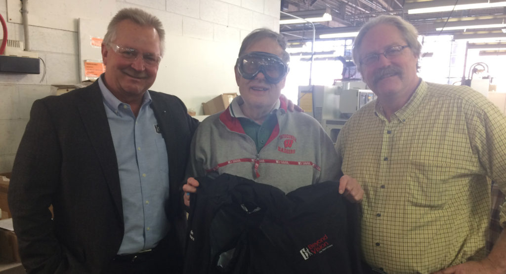 On the left, Jim Kerlin, center is Gale holding a black jacket with his name on it and on the right is Ron Howski. They are all smiling.