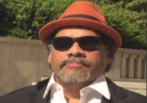 A man with sunglasses, a goatee and a red fedora is standing outside. He is wearing a dark suit, white dress shirt and has a red handkerchief in his breast pocket.