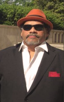A man with sunglasses, a goatee and a red fedora is standing outside. He is wearing a dark suit, white dress shirt and has a red handkerchief in his breast pocket.