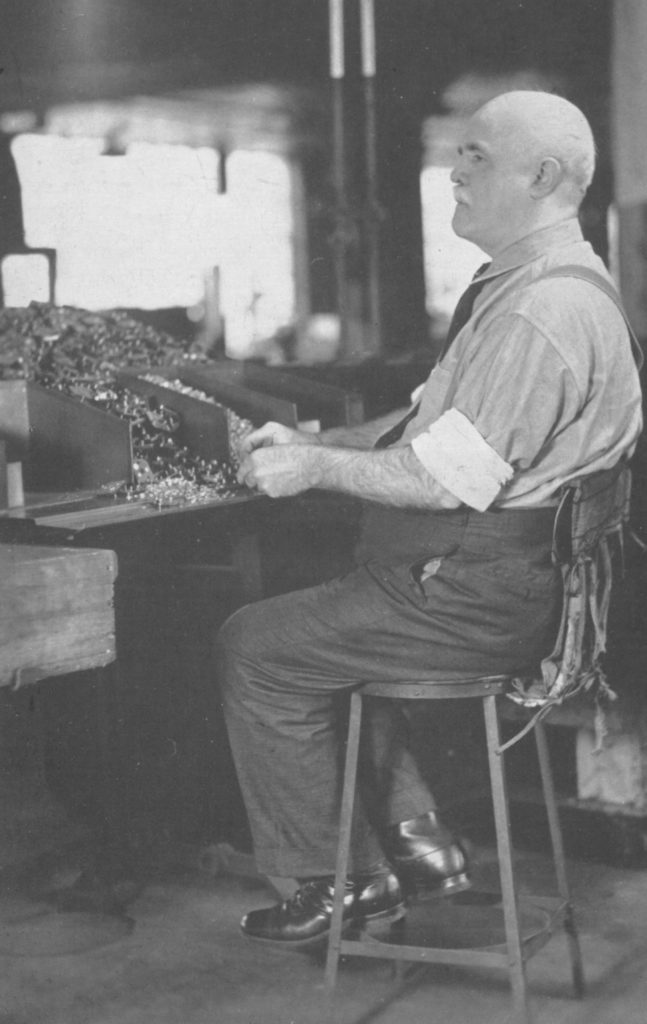 A man in his fifties is sitting at a high stool in front of some bins on a table. He is bald and has a white mustache. His shoes are very shiny black dress shoes and he has on some dress pants, a collared shirt, tie, suspenders and his sleeves rolled up above his elbows. There is a large, wide bin to his left and three other bins with small metal parts flowing out of them and he holding some parts in his hands.