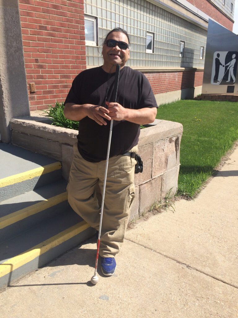 Julius stands in front of the entrance to Beyond Vision. He's wearing khakis and a black tshirt, holding his cane and laughing.