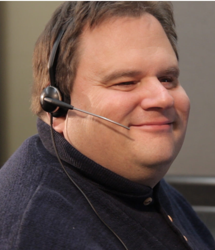 Headshot of Steven Gastreich smiling, wearing a headset at a computer.