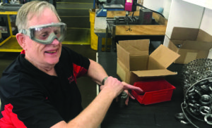 Gale Klatt sits at a work table. He is wearing safety goggles and smiling brightly. He is wearing a black and red Beyond Vision shirt.