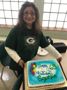 Sophia Kipp stands with a large birthday cake that reads. "Happy 65th Birthday, Sophia!!" She is wearing a Green Bay Packers shirt and smiling!