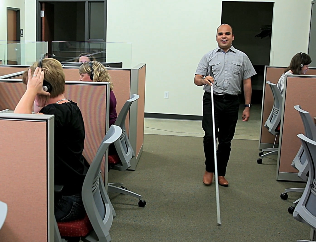 Israel uses a long white cane to walk between a row of cubicles. The people sitting at the desks are wearing headsets.