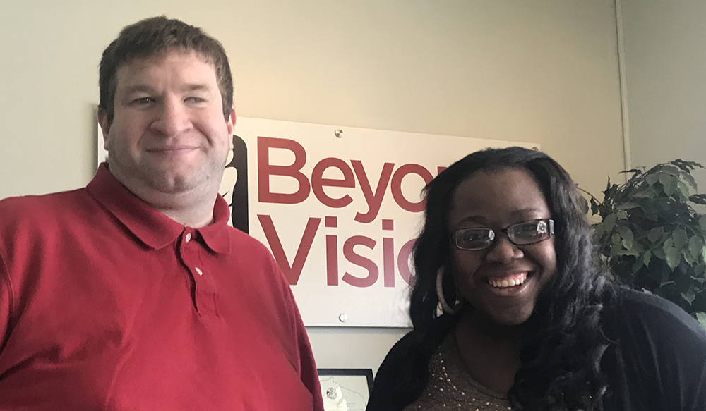 Ruben and DeLonna stand in the lobby by the Beyond Vision sign. He is smiling and wearing a red polo. DeLonna is laughing wearing a gold top with black sweater, glasses and long dark hair.