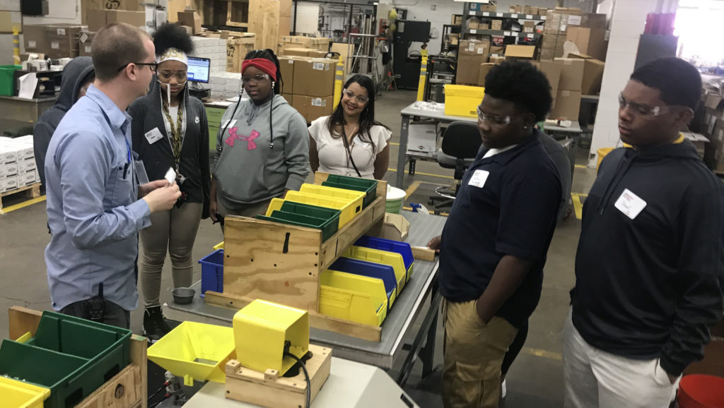 students lean over some assembly parts bins as a young man with glasses shows them how the work is done.