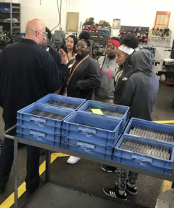 A group of students listen as a tall man talks to them in a machine shop.