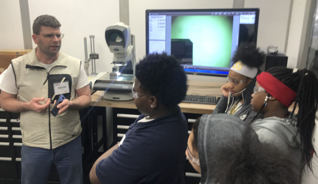 A man shows students how he measures machine parts using lasers and a screen with magnification.