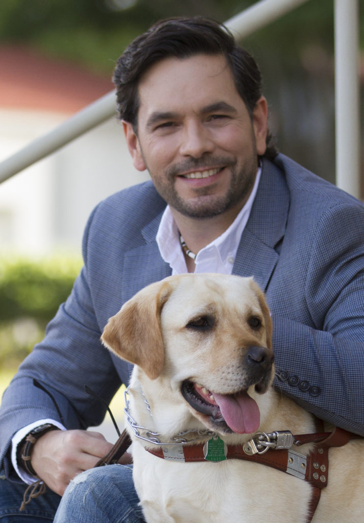 Beyond Vision employee, Guillermo Baena wearing a blue suit and sitting on a step next to his guide dog.