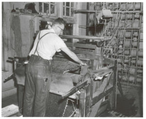 A man in overalls is leaning over a loom that has many spools of thread leading to it