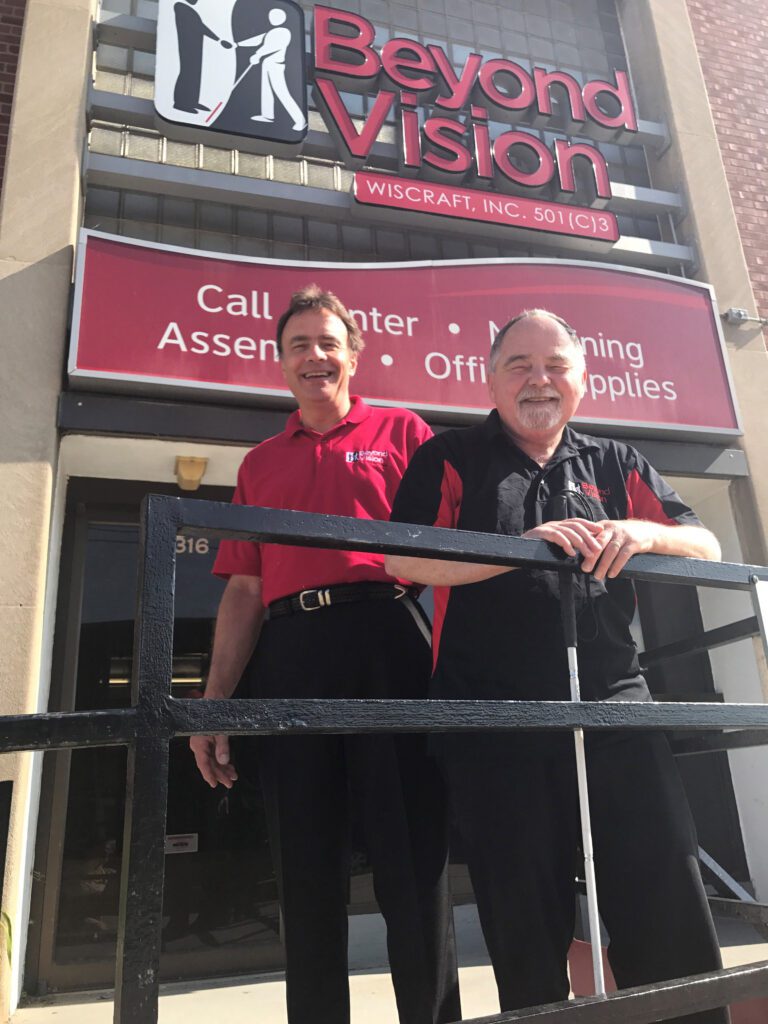 Two men are standing at the entrance of Beyond Vision laughing. One is holding a white cane.