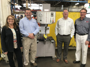 a woman and three men stand next to a narrow machine and smile. They are in manufacturing space.
