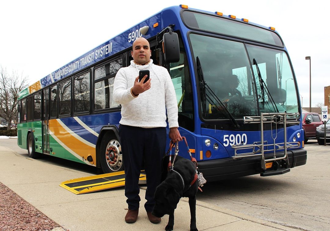 a man with a phone is standing with a guide dog near a city bus.