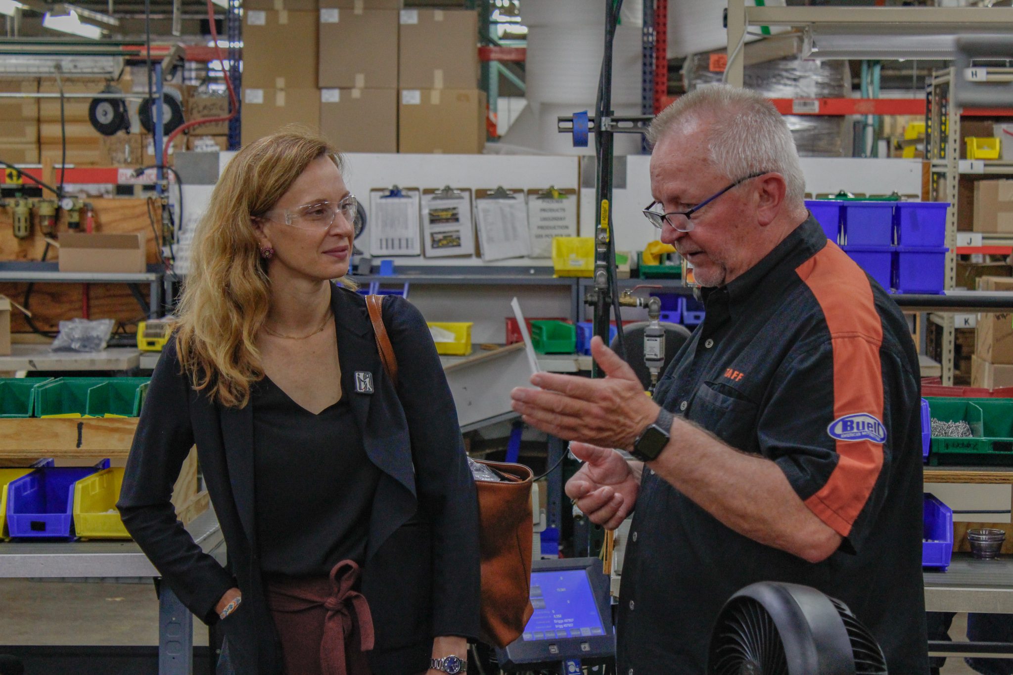 A man and a woman wearing safety glasses are talking in a warehouse space with racks of boxes and workbenches around them.