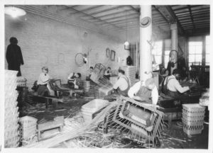 a black and white picture from the 1930s with a room where men in overalls are weaving baskets. At one end of the room there are stacks of baskets that are completed.